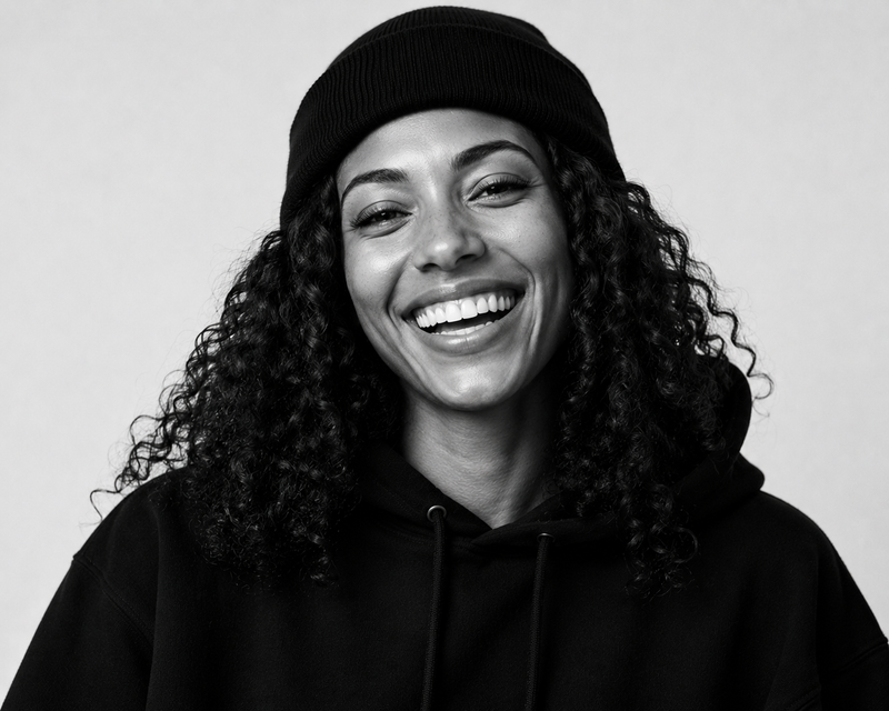 A black and white portrait of a smiling woman with curly hair wearing a black beanie and a black hoodie against a light gray background.